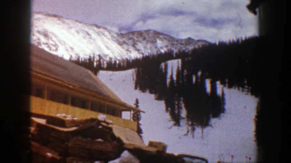 Aspen, Colorado winter view from 1961 with a low rustic building, snowfield, evergreen trees, and pale mountain ridge in the distance.