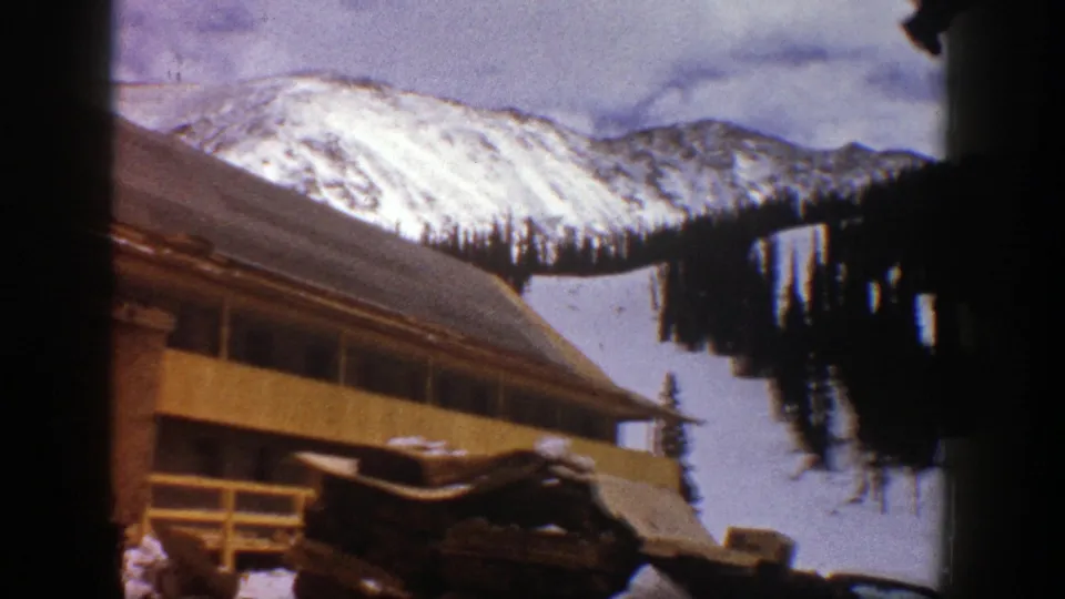1961 Aspen winter frame with the cabin or lodge roof and windows below a snow-covered hillside and evergreen cluster.