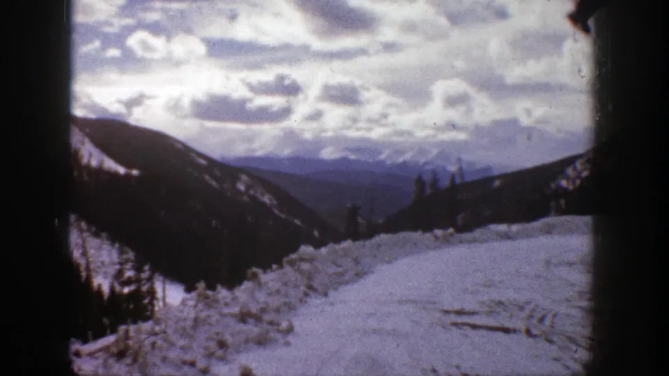 Aspen, Colorado home-movie frame showing a snow-covered overlook, tree-lined slopes, distant blue ridges, and dark framing at the sides.