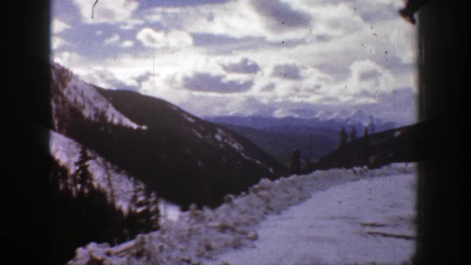 1961 8mm frame from Aspen showing a snowy foreground, dark forested valley walls, distant mountain layers, and bright broken clouds.