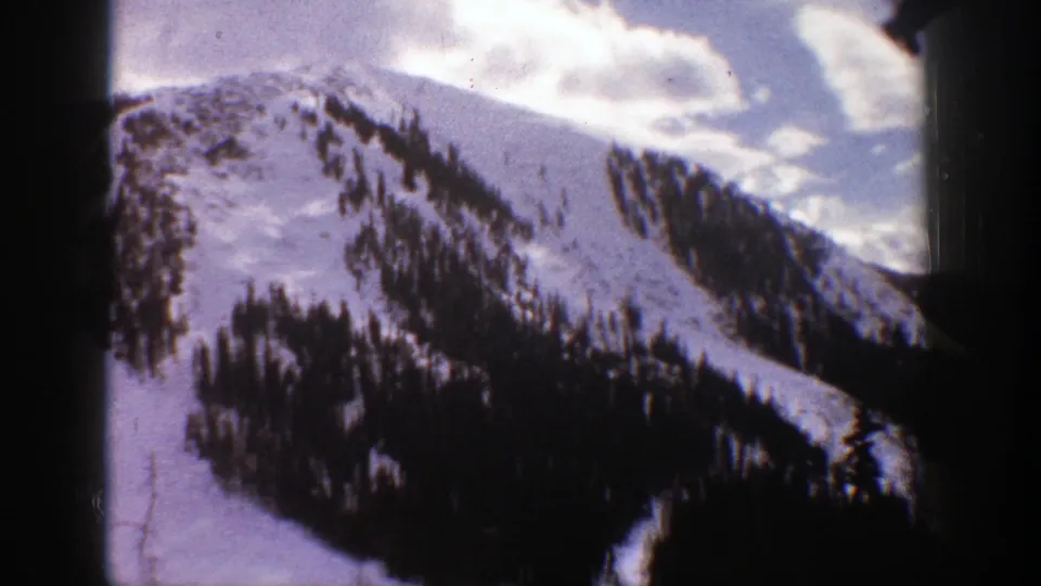 1961 Aspen frame showing a broad snow-covered mountainside, irregular evergreen patches, and bright clouded sky at the ridge line.