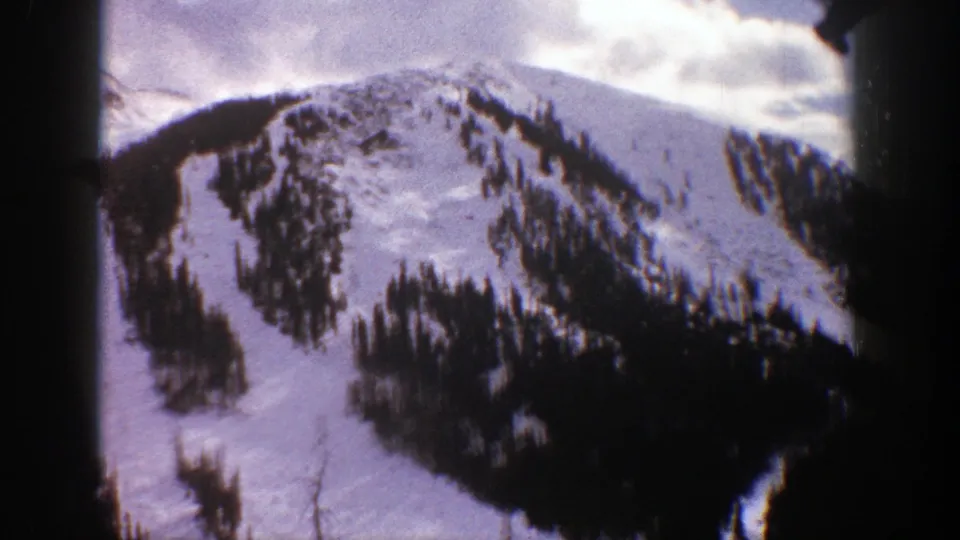 1961 Aspen home-movie view of a snow-covered mountain face with scattered evergreens, a dark lower slope, and bright clouds overhead.