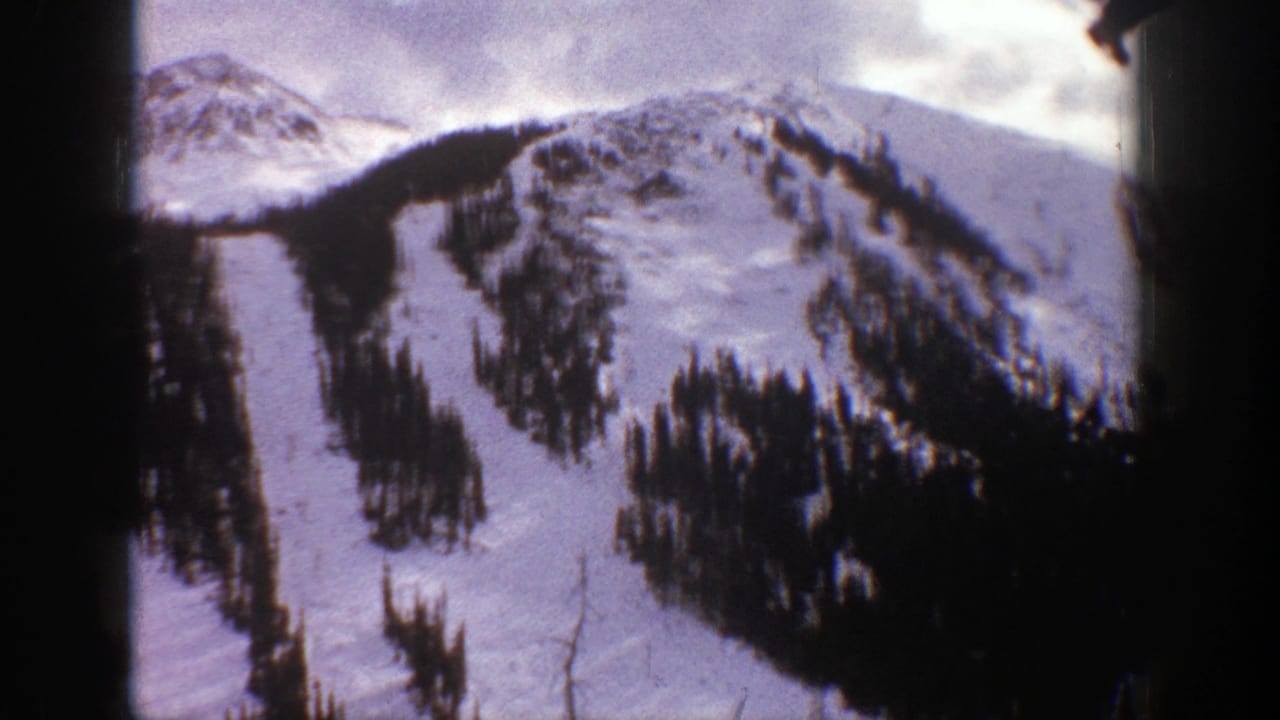 Snowy ski runs descend through dark conifer trees below rocky mountain peaks.