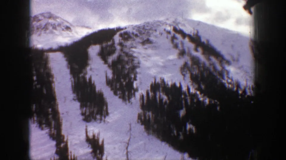1961 Aspen, Colorado home-movie frame showing snow-covered ski runs bordered by conifers beneath cloudy mountain ridges.
