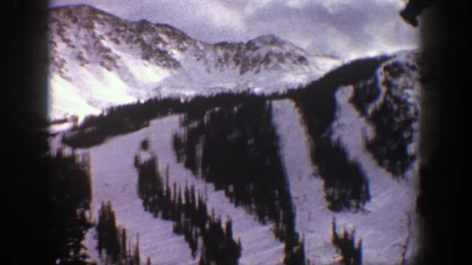 1961 8mm view of Aspen ski slopes, showing snow-covered runs, dark tree lines, and rugged peaks under cloud cover.