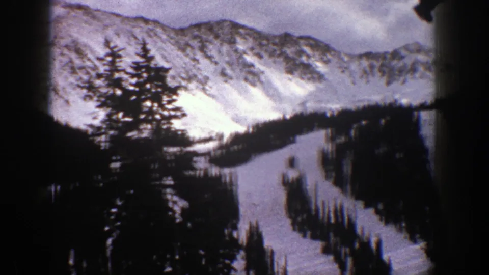 1961 Aspen, Colorado 8mm frame showing snowy peaks, dark conifers, and a winding white slope through the trees.