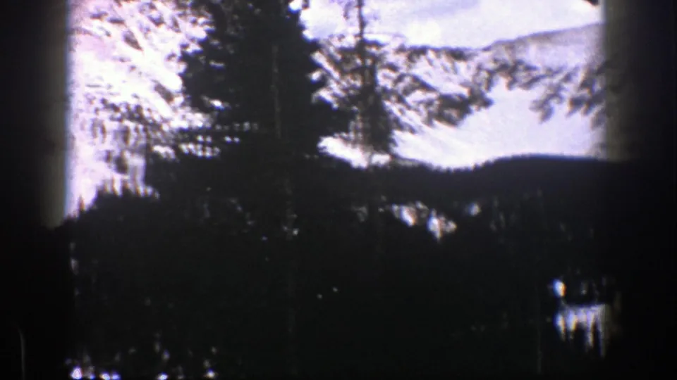 1961 Aspen home-movie frame showing snow-covered mountains beyond a shadowed evergreen forest, with soft grain and film gate edges.