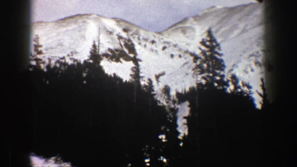 Aspen 8mm home-movie frame with bright snowy peaks behind a dense dark evergreen foreground and visible film texture.