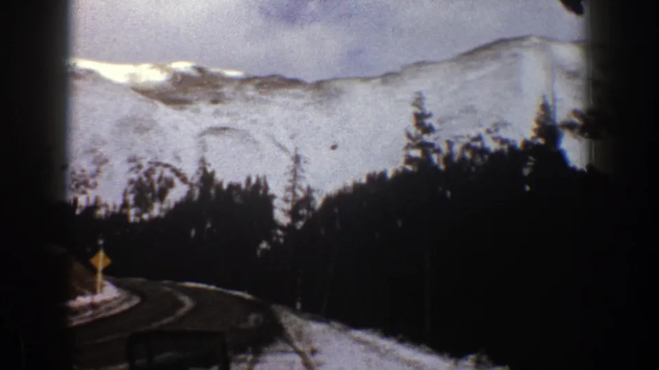 1961 Aspen, Colorado 8mm view of a winding road, roadside snow, evergreens, and snowy mountain slopes.