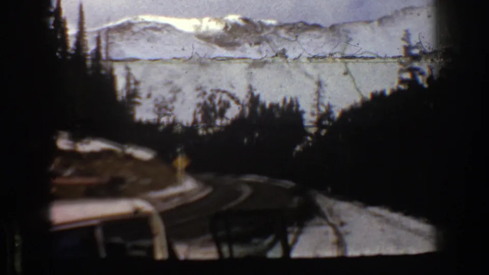 1961 Aspen, Colorado frame with a partial vehicle foreground, winding mountain road, evergreens, and snowy ridges.