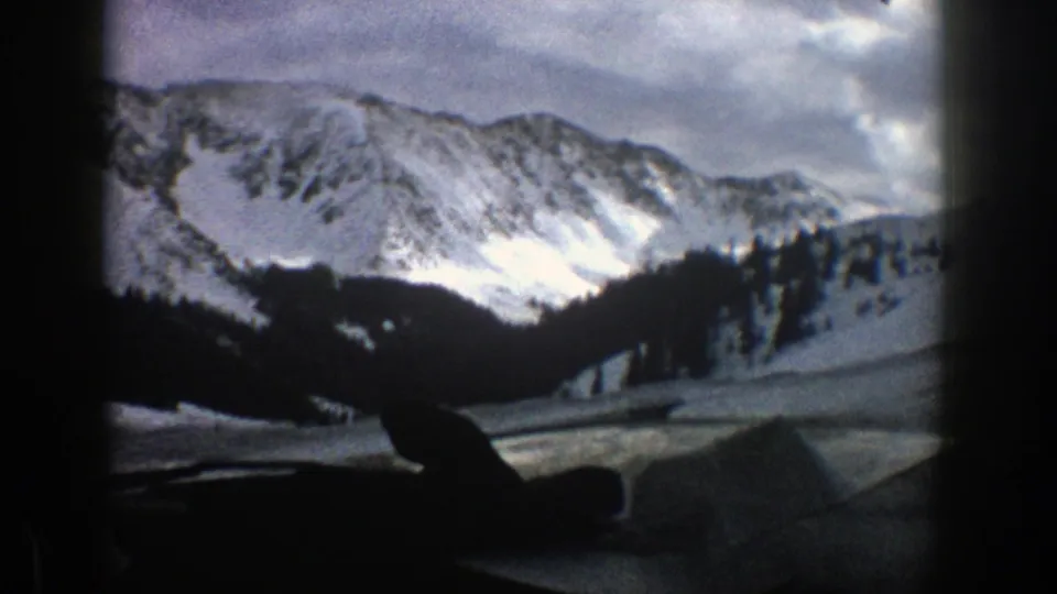 1961 Aspen-area winter mountains viewed from inside a moving vehicle, with snowy slopes, dark trees, and soft 8mm grain.