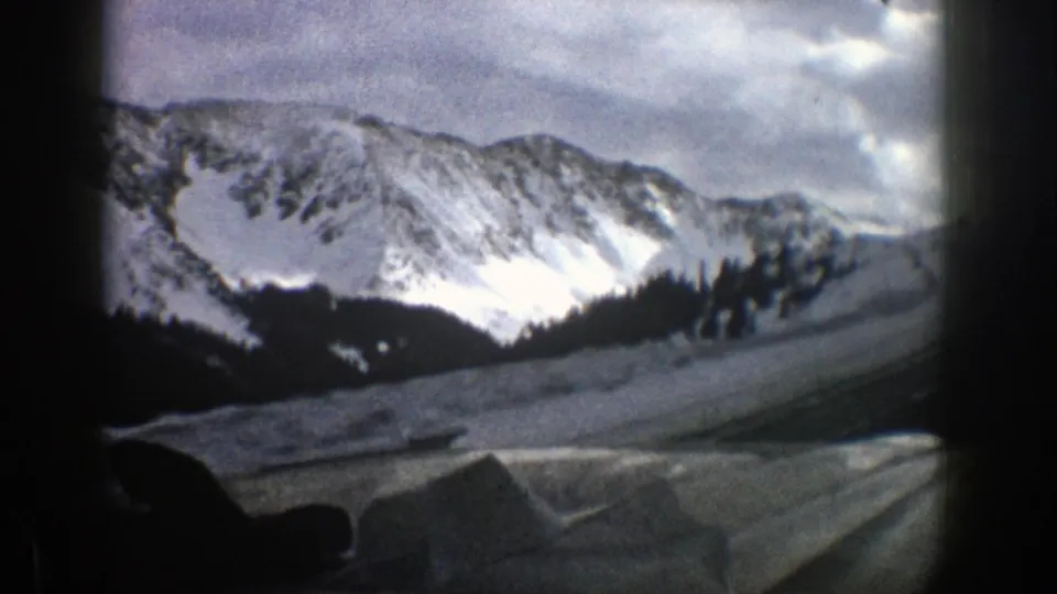 1961 Aspen winter view with snow-covered mountains, a dark wooded ridge, and a curving road or snowbank seen through a vehicle window.