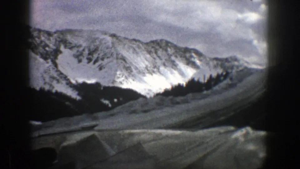 Aspen winter mountain scene on 8mm film, with a snowy ridge, evergreen trees, and a pale road or slope below the vehicle window.