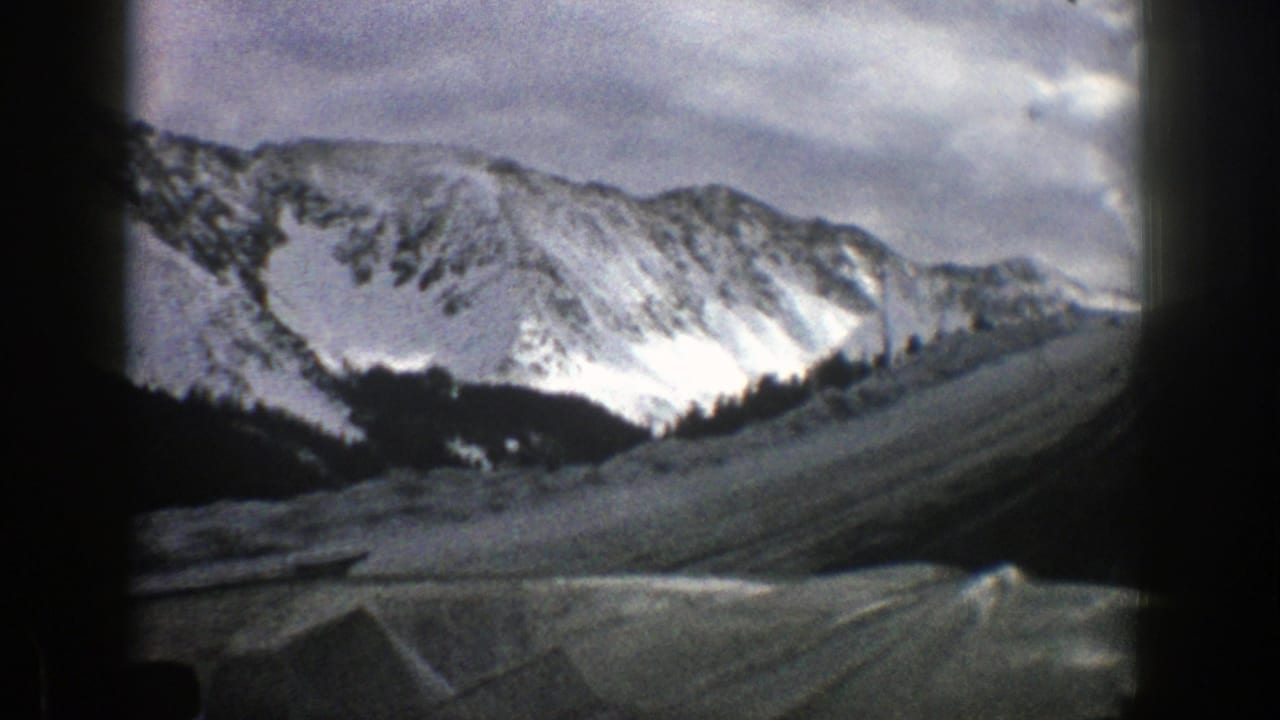 Snowy mountain road seen through a car windshield with peaks under a cloudy sky.