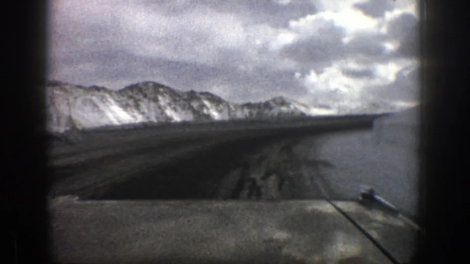 Home-movie frame from a 1961 Aspen drive, with the car hood below a snowy road and mountain skyline.