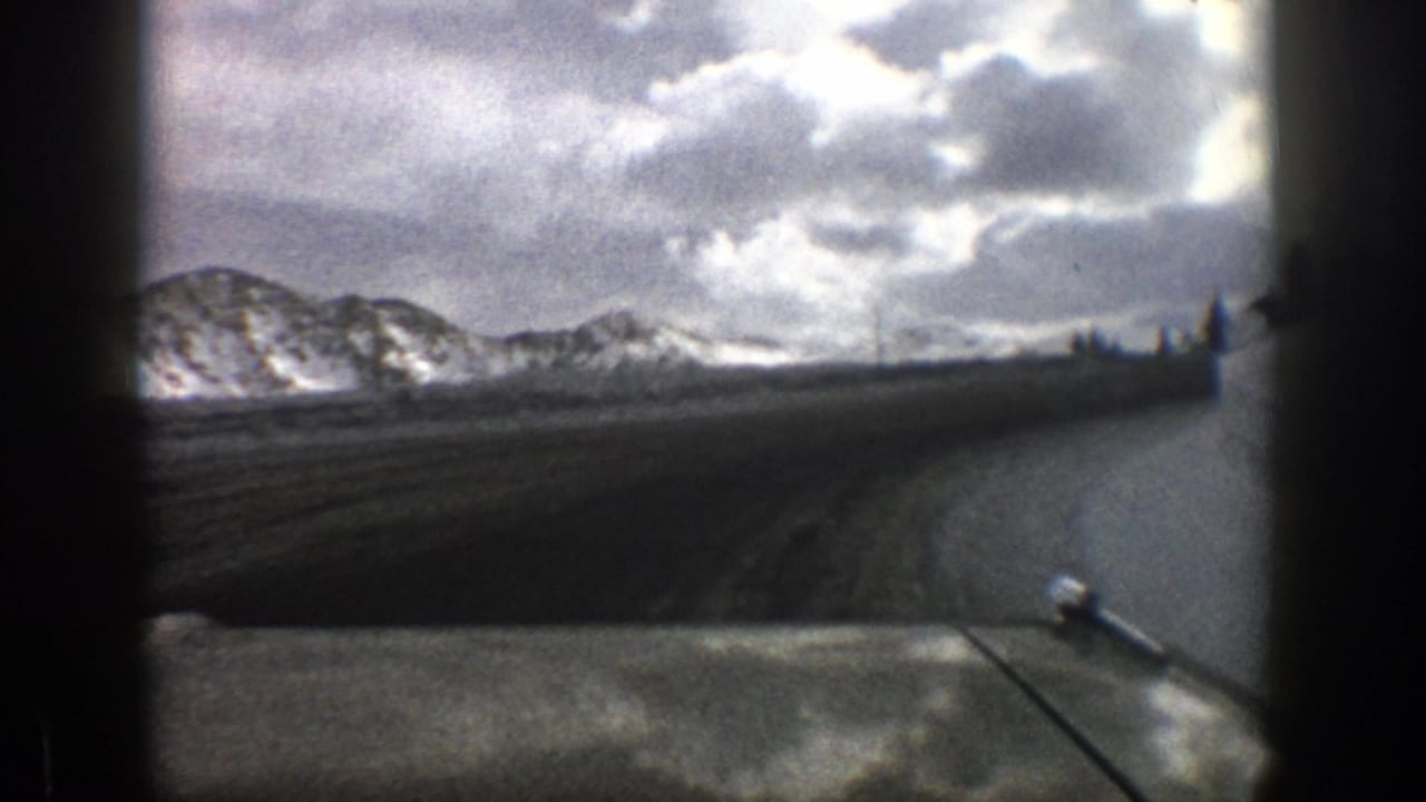 1961 Aspen-area home-movie frame showing a road curving toward snow-covered mountains beneath a stormy sky.