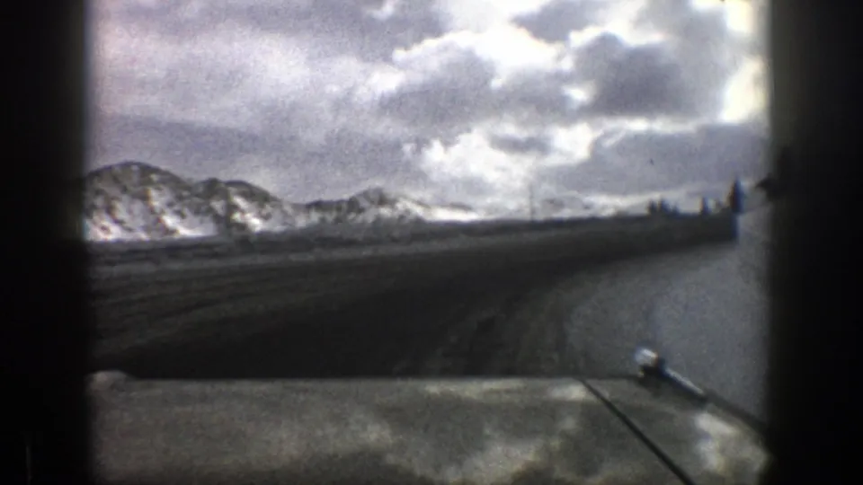 1961 Aspen-area home-movie frame showing a road curving toward snow-covered mountains beneath a stormy sky.