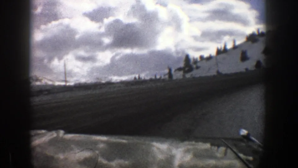 A mid-century 8mm travel view follows a curving snowy road near Aspen, with a tall roadside pole and clouded mountain backdrop.