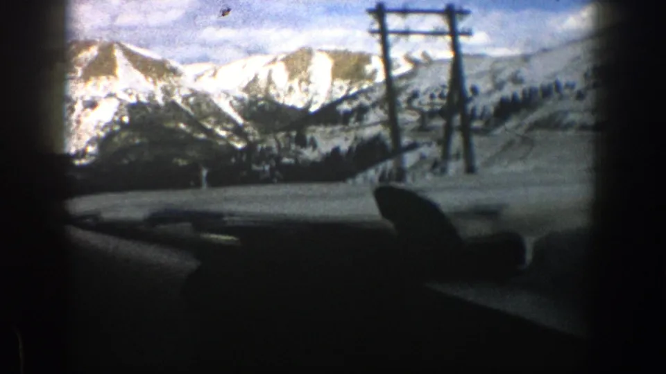 A 1961 Aspen-area car ride passes utility poles with snow-covered mountains and trees beyond the windshield.