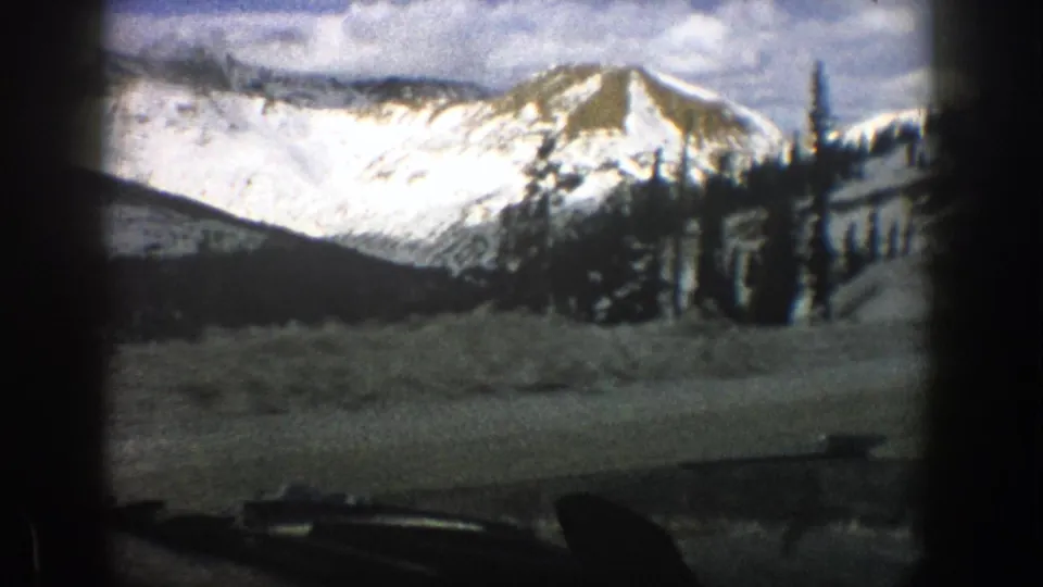 1961 Aspen, Colorado roadside view with snowbanks, dark trees, and a sunlit mountain slope beyond the windshield.