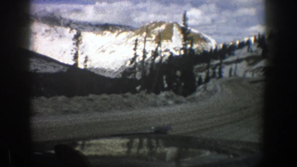 1961 Aspen, Colorado home-movie frame showing a snowy roadside curve, evergreens, and mountains through the windshield.