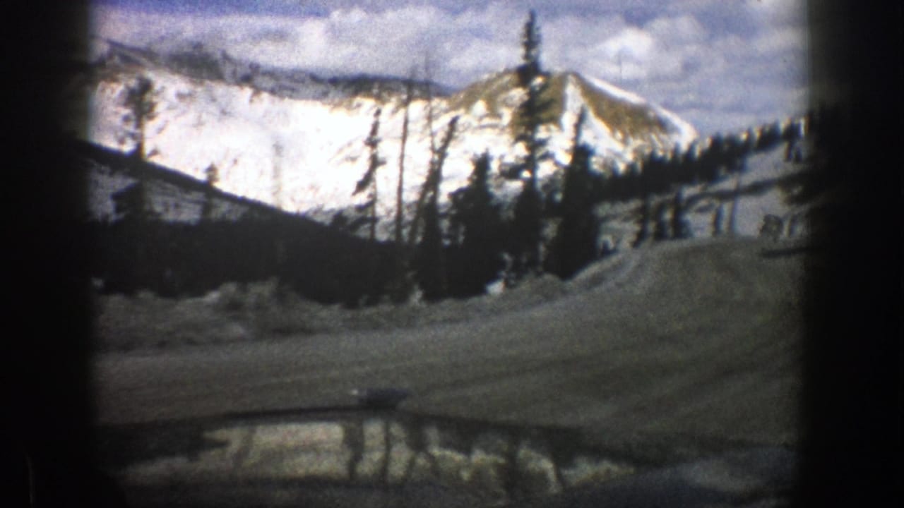 Snowy mountain road with evergreen trees and distant peaks under a cloudy sky.
