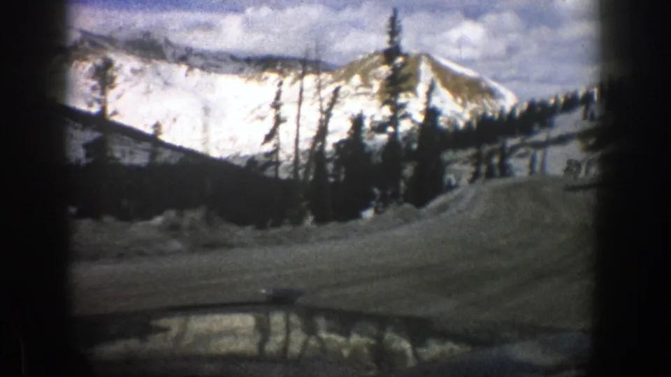 1961 Aspen-area 8mm frame showing a snowy road, dark evergreens, and snow-covered mountain slopes ahead.