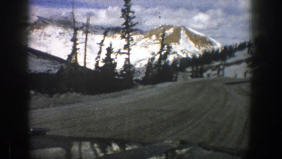 Moving 8mm view of a snowy Aspen mountain road curving past evergreens with bright slopes in the distance.
