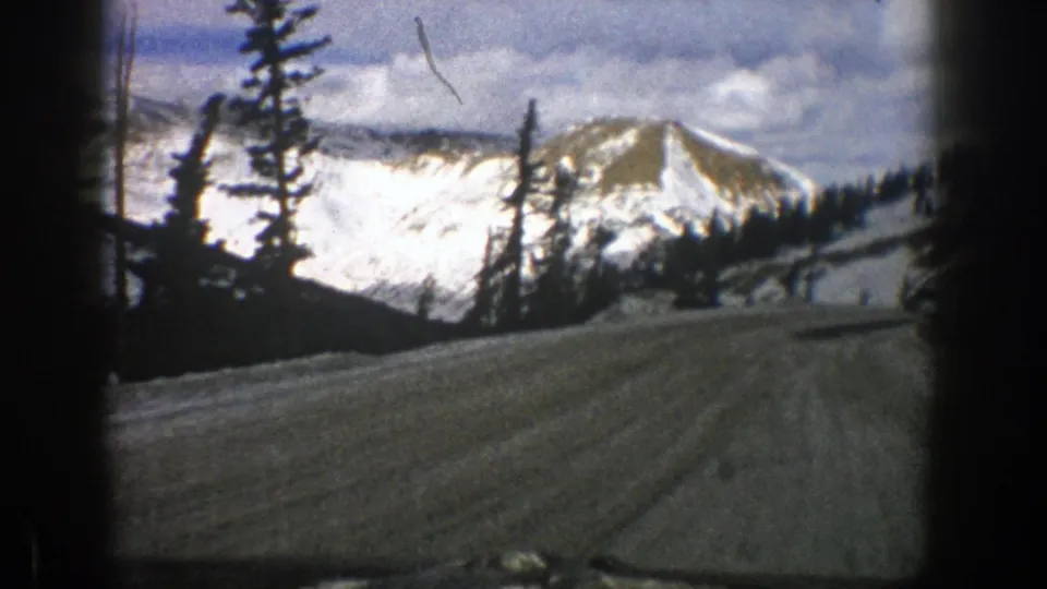 1961 home-movie frame from Aspen showing a rough snowy road below tall evergreens and a bright mountain ridge.