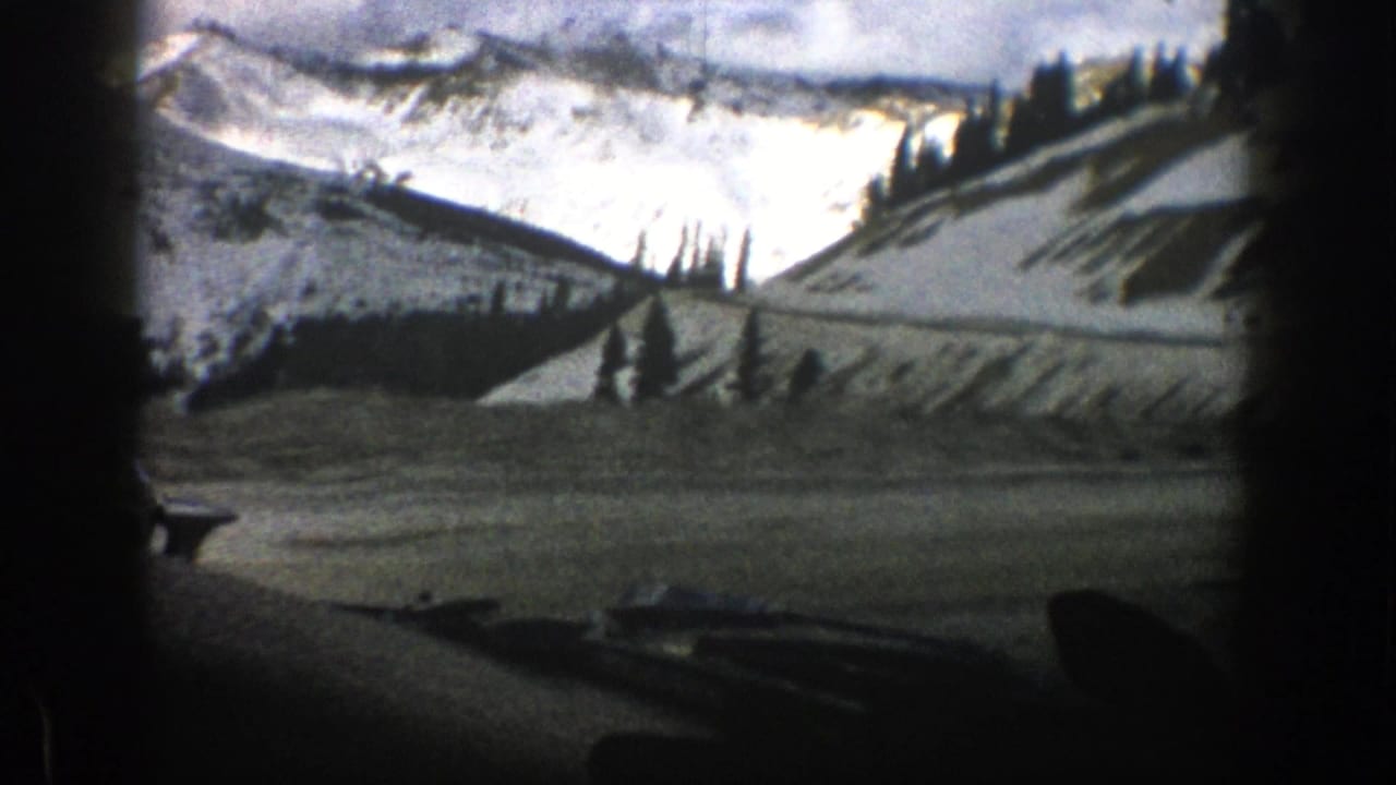 Windshield view of a snowy mountain road with dashboard silhouette and distant peaks.