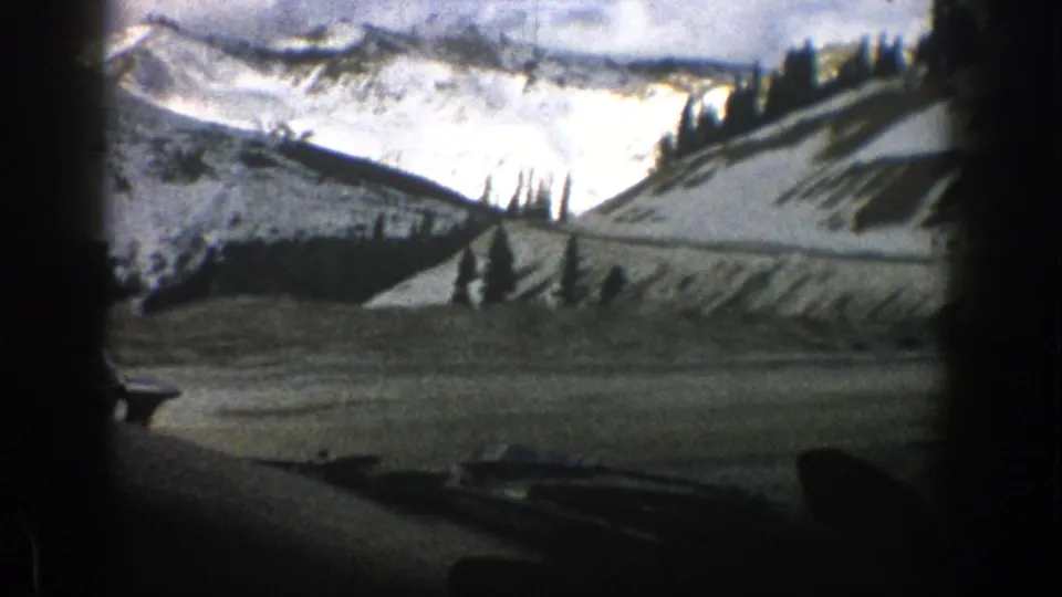 1961 8mm view from inside a vehicle near Aspen, Colorado, looking toward a snowy road, evergreen trees, and distant mountains.
