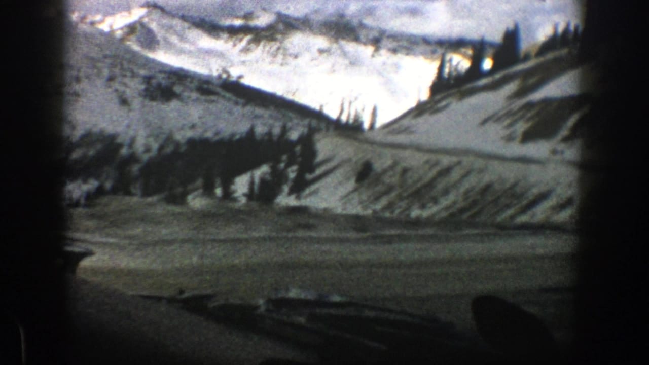 Snowy mountain slope and road seen through a dark-framed vehicle windshield.