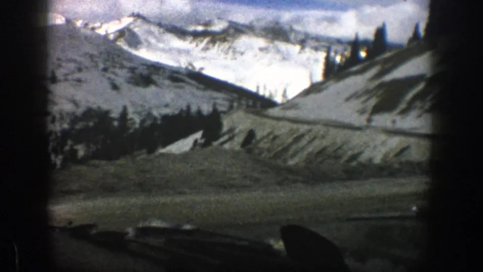 1961 home-movie frame from a moving vehicle near Aspen, Colorado, with dashboard foreground, snowy hillside, road, evergreens, and mountain peaks.