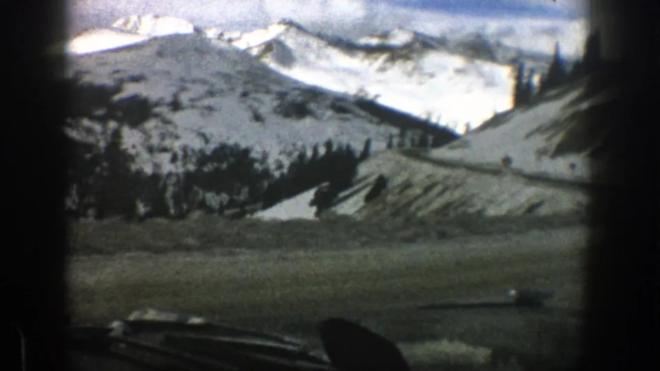 Aspen, Colorado 8mm travel frame from 1961 showing a car windshield view of snowy slopes, evergreen trees, a road, and bright mountain ridges.