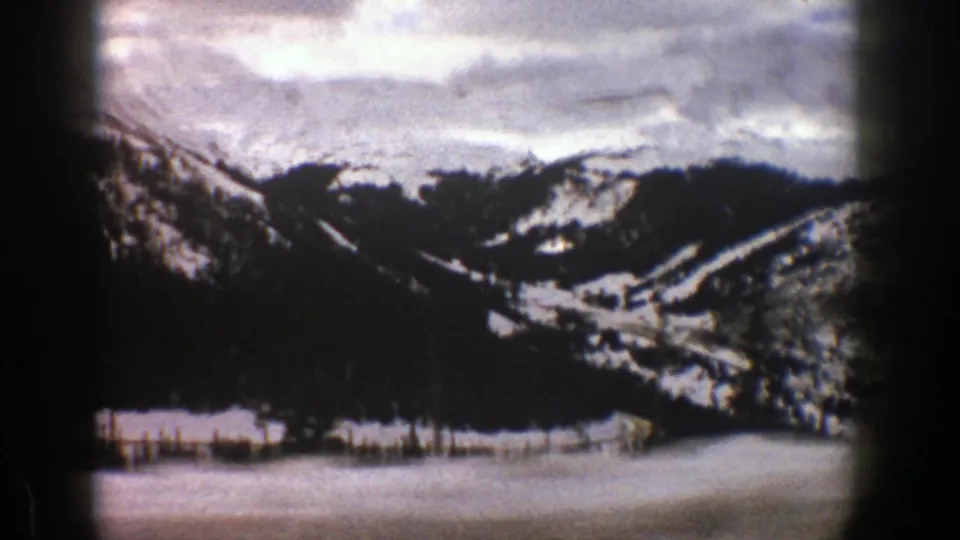 1961 home-movie view near Aspen showing a snowfield in the foreground, dark evergreens, and rugged mountain slopes under a cloudy sky.