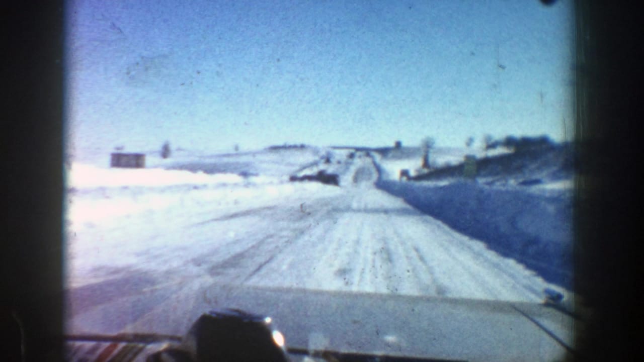 Snow-packed road seen over a car hood with open fields and snowbanks.