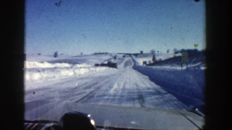 1961 winter driving view from inside or just behind a car, looking along a snow-covered Aspen road under a blue sky.
