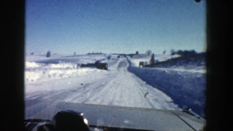 Home-movie frame from an Aspen winter drive, showing a snow-packed road, car hood, and open roadside snowfields.