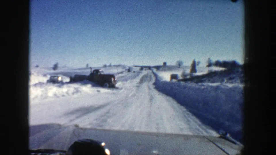Interior vehicle view on a 1961 Aspen snowy road, with tracks ahead and a parked truck near the left shoulder.