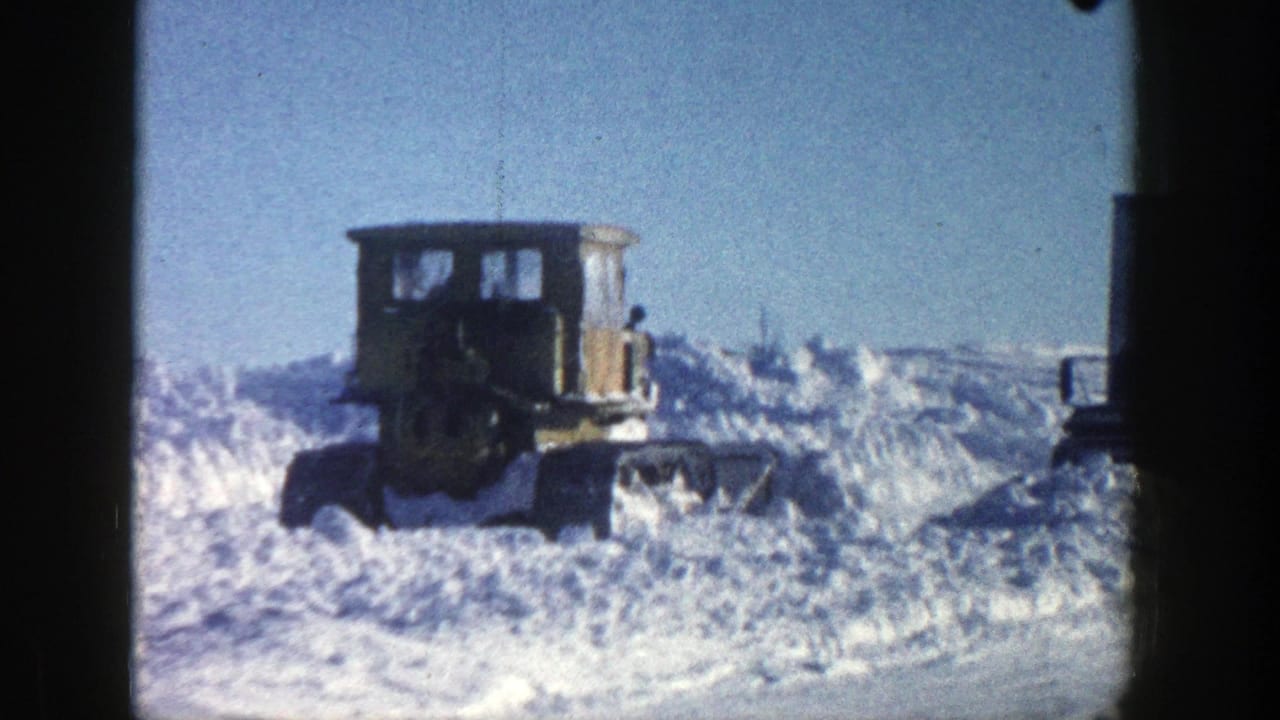 1961 Aspen, Colorado 8mm frame showing a yellow tracked snow plow clearing deep snow beneath a bright blue sky.