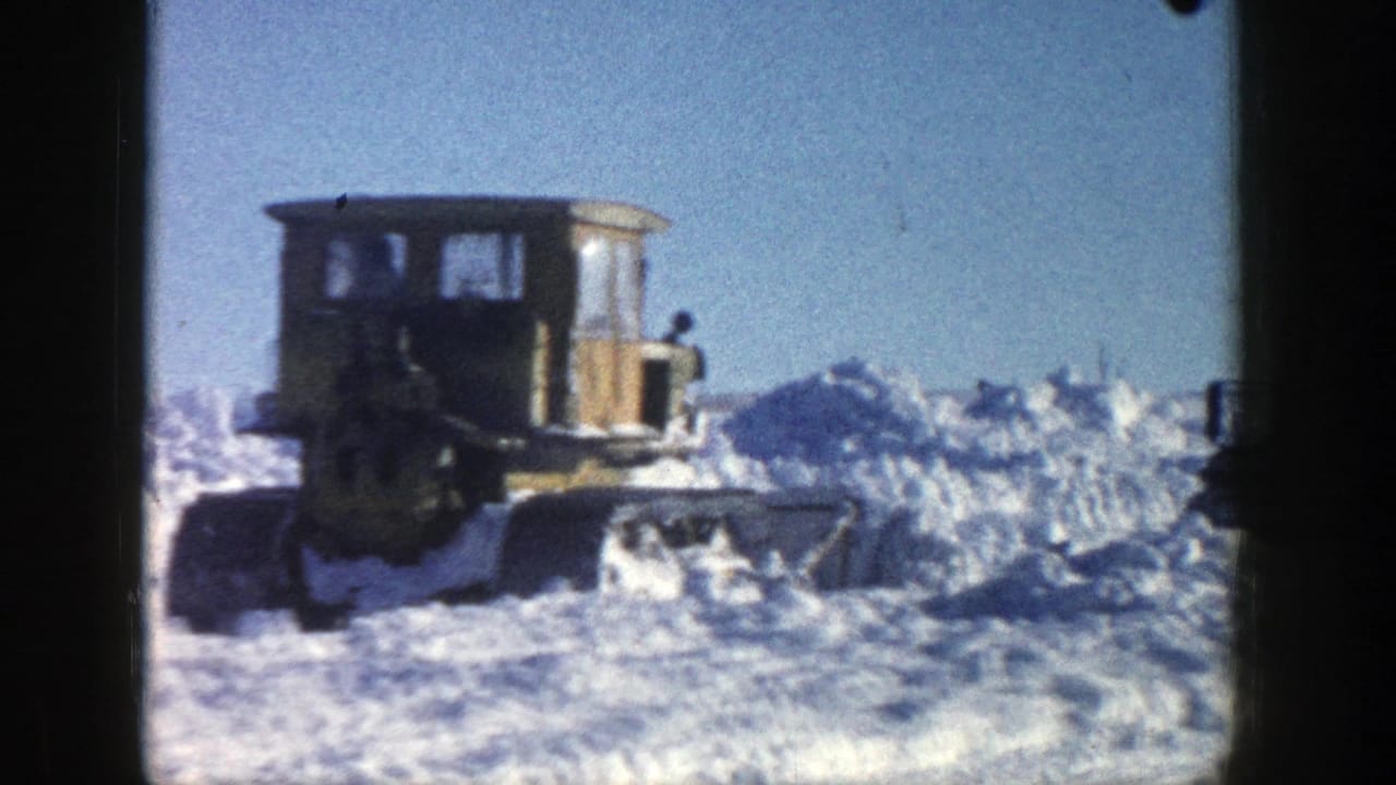 Tracked snow plow advancing through snow with the cab and blade visible.