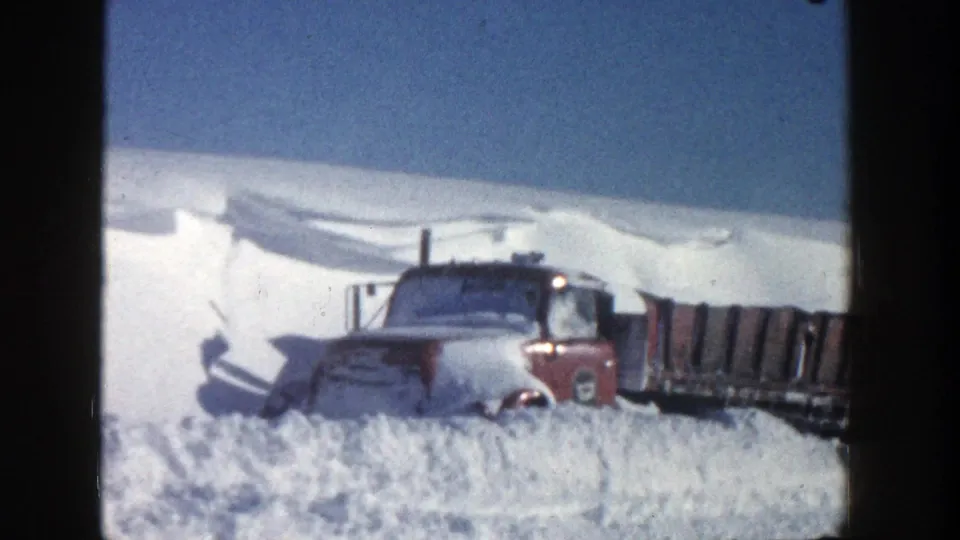 Closer 8mm frame of the red truck cab in deep snow, with the long bed extending to the right under a clear sky.