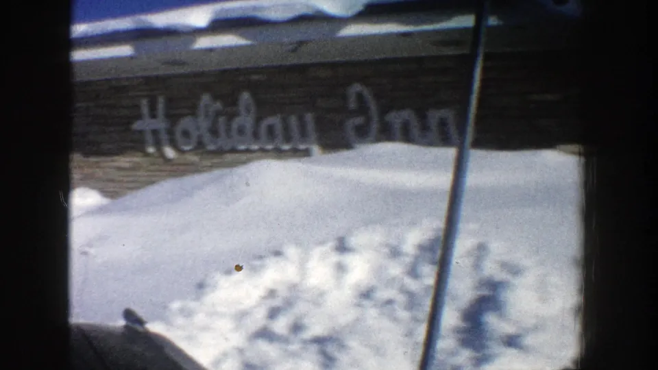 1961 Aspen winter drive-by frame showing a Holiday Inn sign partly obscured by high snowbanks and dark vehicle framing.
