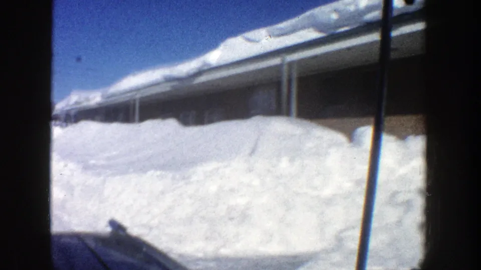 1961 Aspen vehicle-perspective view of a low building, snow-loaded roof, and packed snow piled along the roadside.