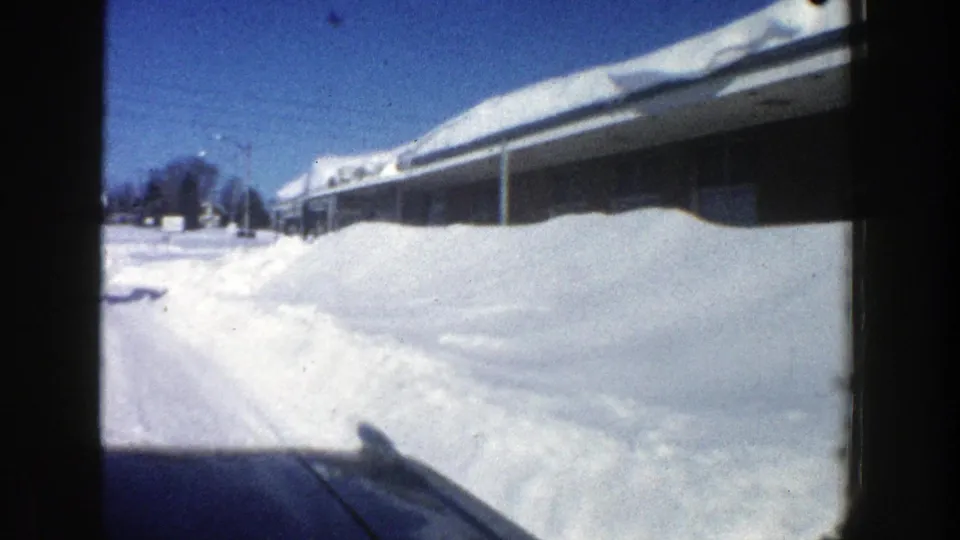 1961 Aspen drive-by frame showing a snow-packed roadside, long low lodging building, roof snow, and distant winter trees.
