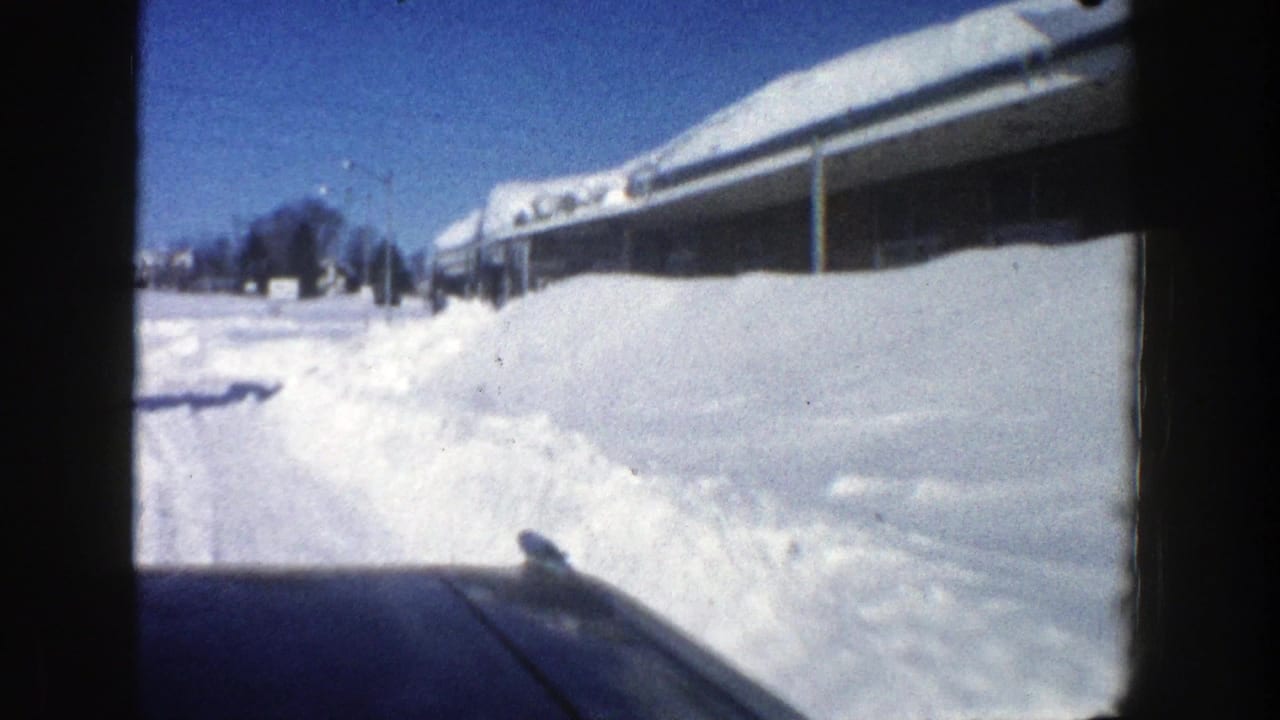 Passenger-view 8mm frame of a snowy Aspen road in 1961, with a car hood, high snowbanks, and a roof buried in snow.