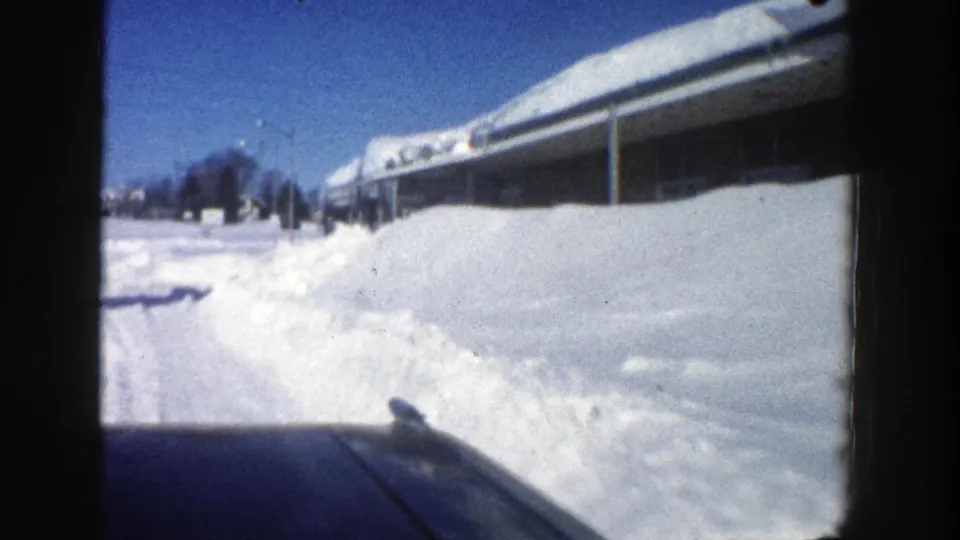 Passenger-view 8mm frame of a snowy Aspen road in 1961, with a car hood, high snowbanks, and a roof buried in snow.