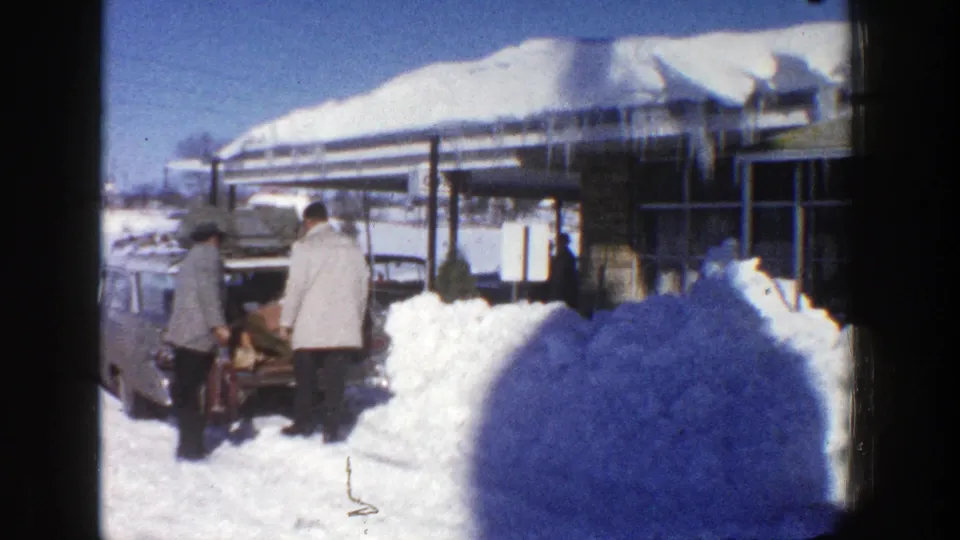 Two men in winter coats unload items from an open trunk near a snow-covered building in Aspen, Colorado, in 1961.