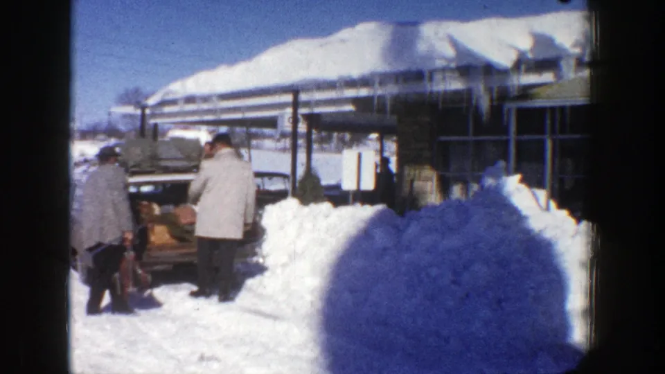 1961 Aspen home-movie frame showing two men at an open car trunk with boxes or luggage amid deep snowbanks.