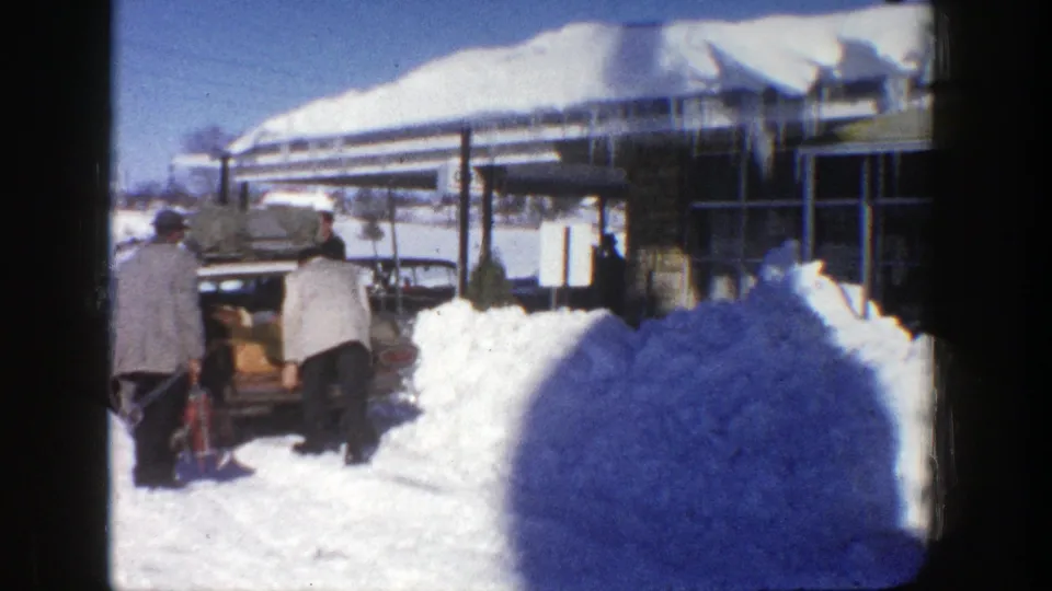 Snowy Aspen arrival scene from 1961, with one man leaning into a vintage car trunk and another standing beside him.